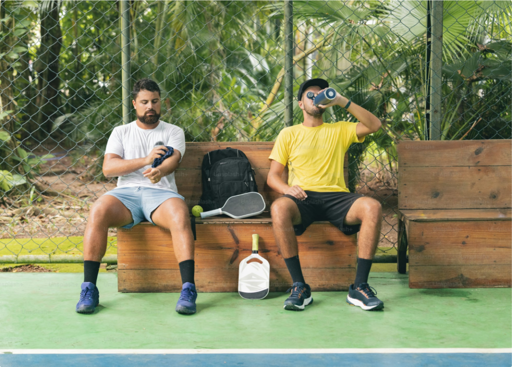 Two men taking a break on a padel court, sitting on wooden benches. One is wearing a white shirt and wiping sweat, while the other, in a yellow shirt, drinks water. A padel racket, a ball, and a backpack are placed between them, surrounded by a green outdoor setting.