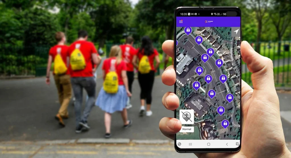 A person holding a smartphone displaying a scavenger hunt map with multiple locked checkpoints. In the background, a group wearing red shirts and yellow backpacks walks through a park, participating in an outdoor scavenger hunt.