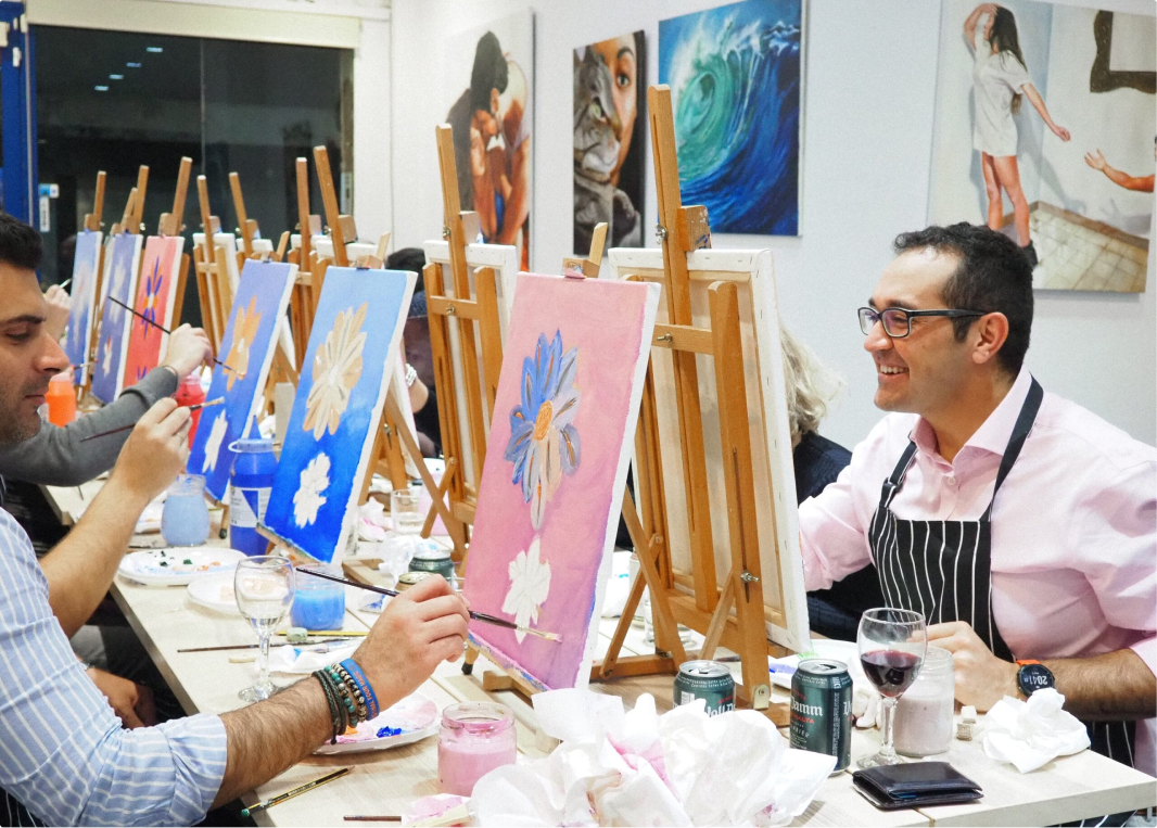 A group of people enjoying a painting class while sipping wine. They are seated at a long table with easels, brushes, and canvases, creating colorful floral artwork in a lively and relaxed atmosphere.