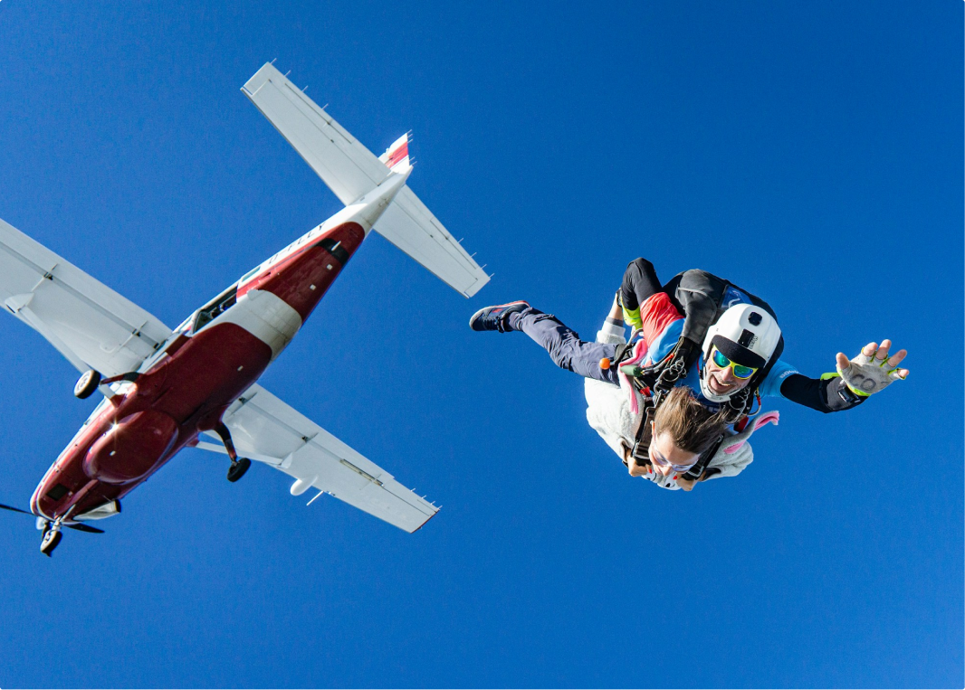 Two people in a tandem skydive, free-falling from an airplane against a clear blue sky. The instructor wears a helmet and goggles, while the passenger extends their arms with excitement. The red and white airplane is visible above them.