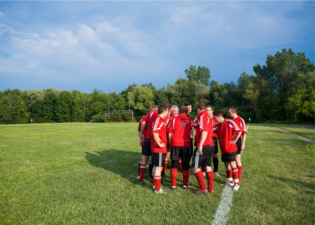 A soccer team in red jerseys huddles together on a grassy field, strategizing before the match. The field is surrounded by trees, with a goalpost in the background.