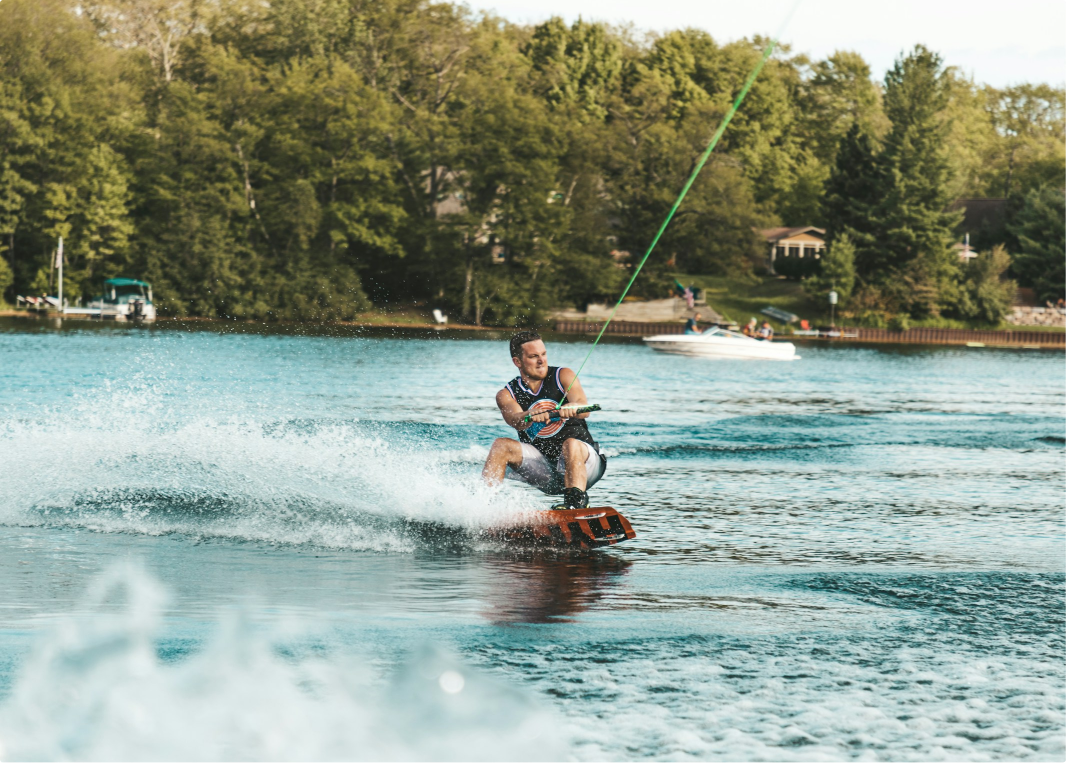 A man wakeboarding on a lake, gripping the tow rope tightly as he carves through the water, creating a spray. He is wearing a life vest, and the lake is surrounded by lush green trees with boats and houses in the background.