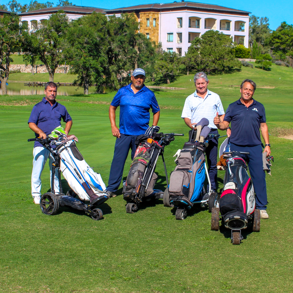 Four golfers pose with their golf trolleys on the course, with a hotel building in the background.