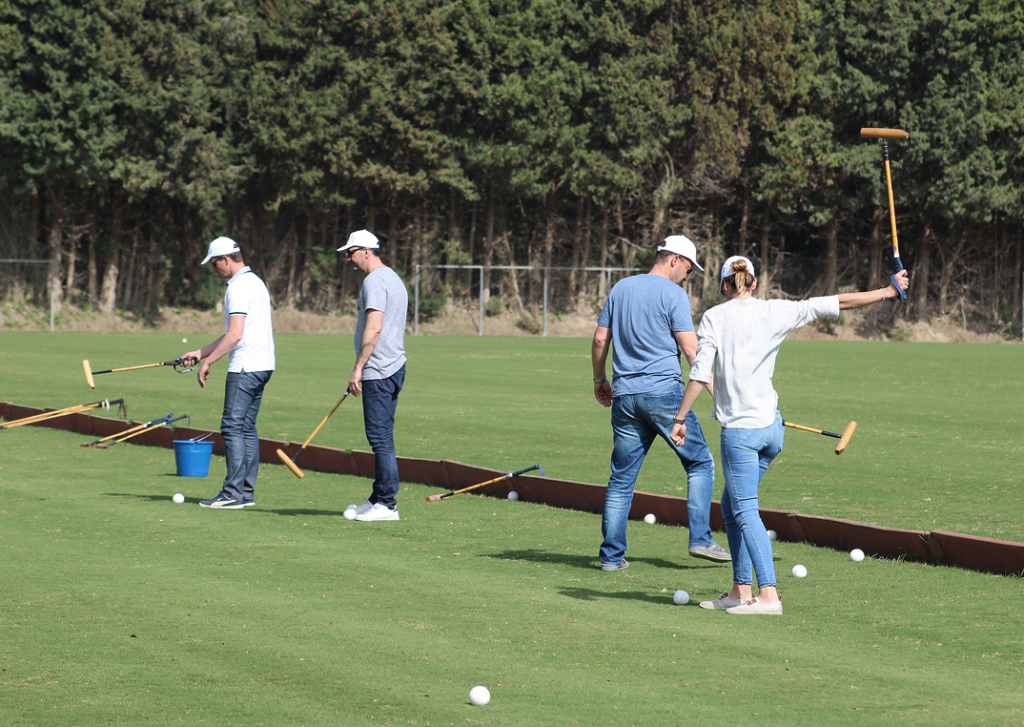 A group of people practicing polo shots on a green field, wearing casual clothes and caps, each holding a polo mallet.