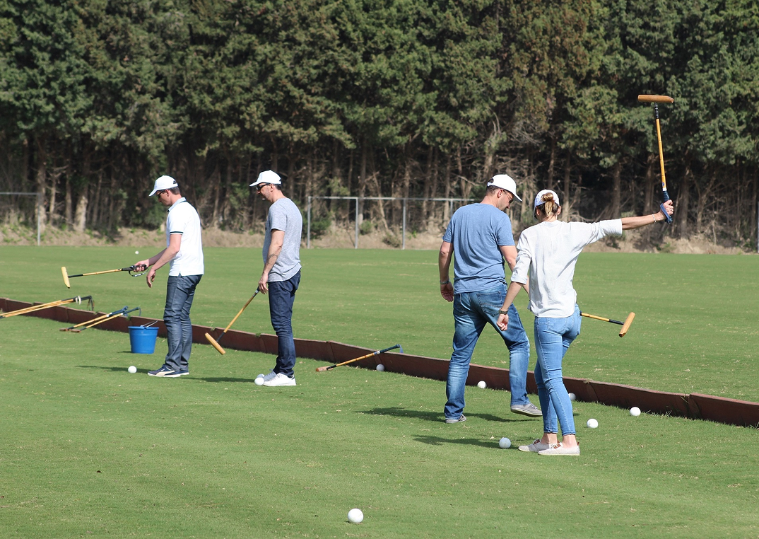 A group of people practicing polo shots on a green field, wearing casual clothes and caps, each holding a polo mallet.